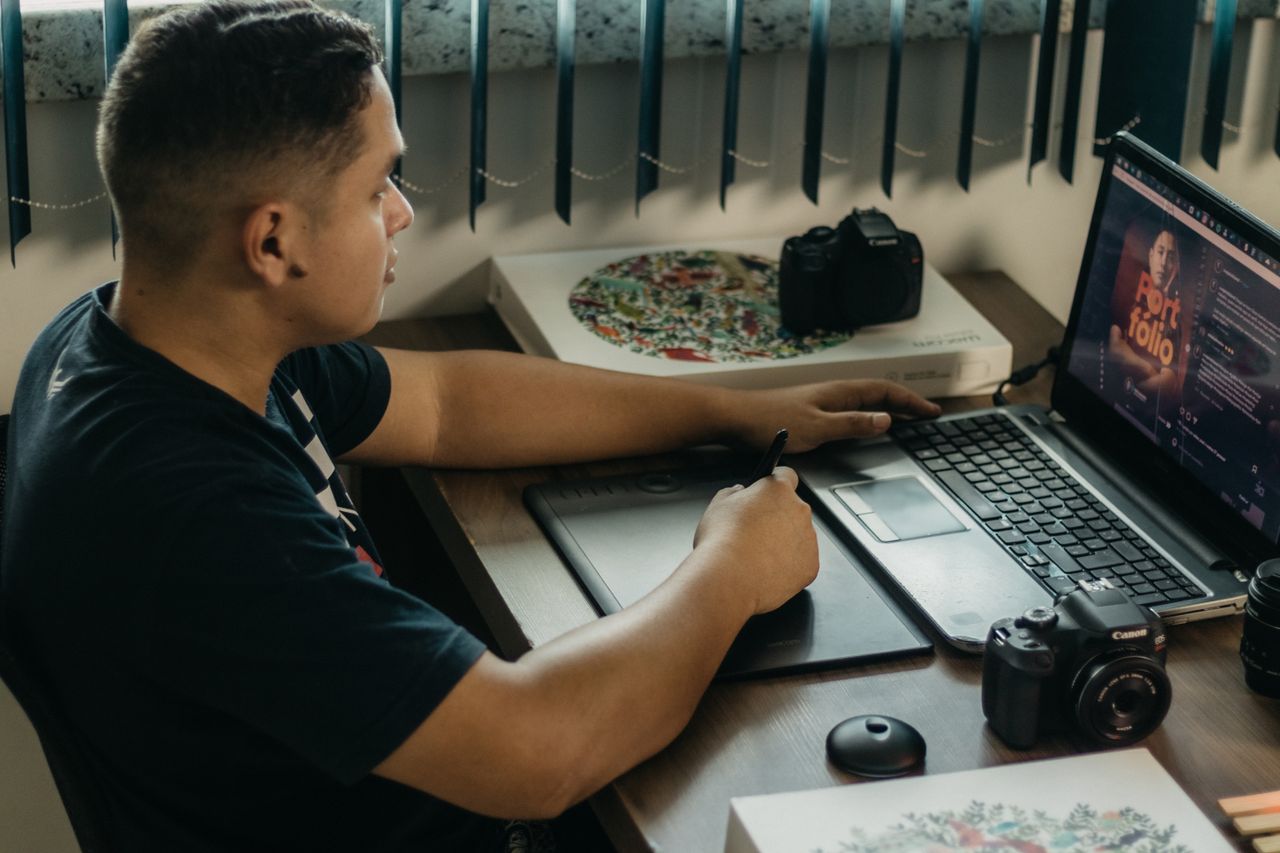 Un homme retouche des photos sur un ordinateur portable à l’aide d’une tablette graphique, dans un espace de travail intérieur avec du matériel photo.